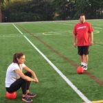 Tom Cavalli standing behind a soccer ball on a soccer field while Alex Morgan sit on a soccer ball across from him.