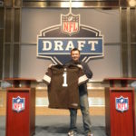Tom Cavalli holding a football jersey between two NFL podiums at the NFL draft