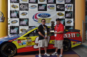 Tom Cavalli and friend holding a trophy in front of a race car at the NASCAR Championship